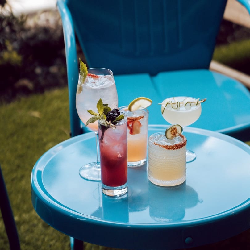 Four colorful cocktails with fruit garnishes on a bright blue outdoor table at Highline Park.