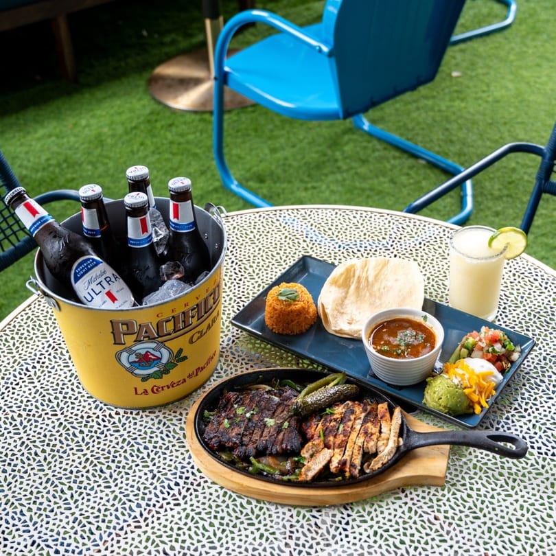Highline Park's fajita platter, beer bucket, and margarita on a patio table with green turf.