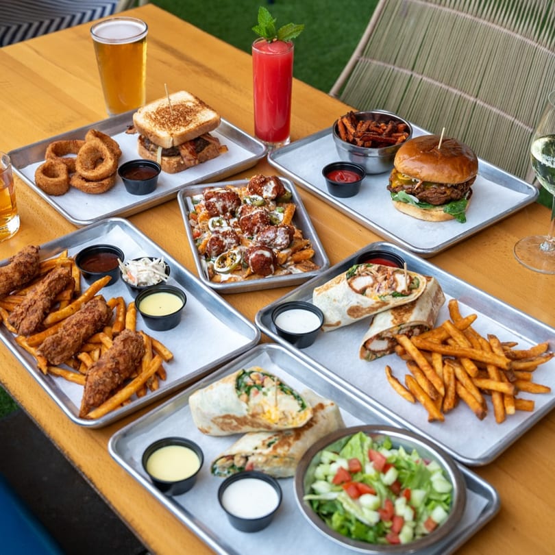 Highline Park bar-&-grill food spread with burgers, wraps, fries, onion rings, and drinks on a wooden table.