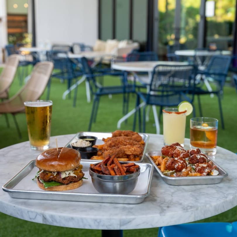 Highline Park patio table with a burger, fries, chicken tenders, and drinks on a sunny day.