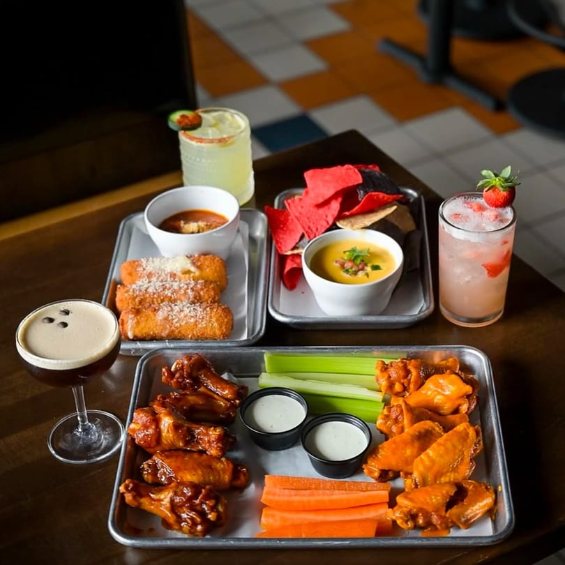 Highline Park bar-and-grill appetizers: wings, mozzarella sticks, chips with queso, and cocktails on a table.