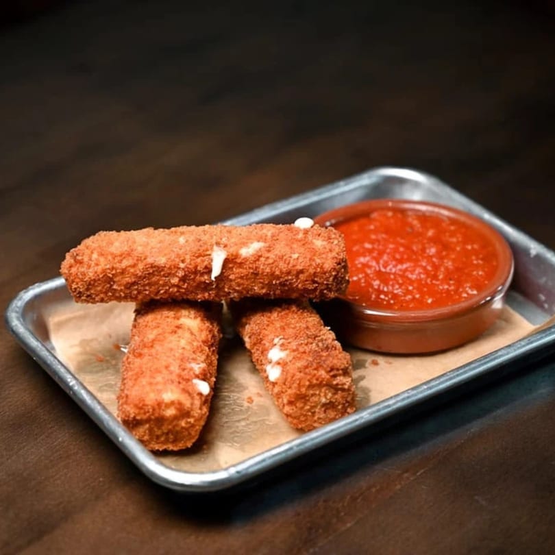 Three crispy mozzarella sticks with melted cheese and a side of red marinara sauce on a metal tray.