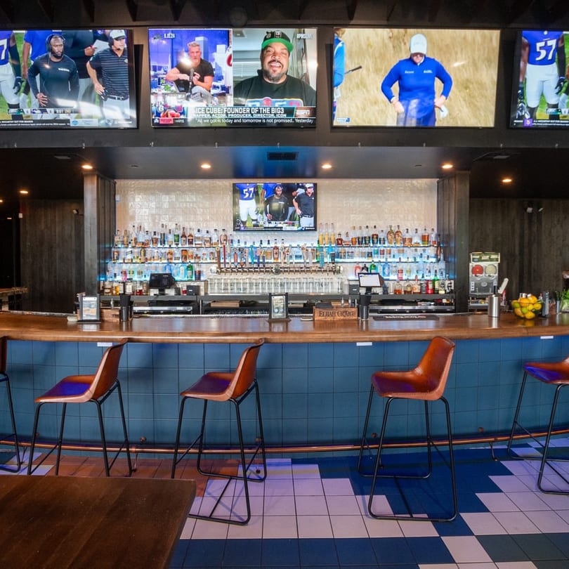 A long bar with brown stools, a tiled blue and white floor, and multiple TVs showing sports.