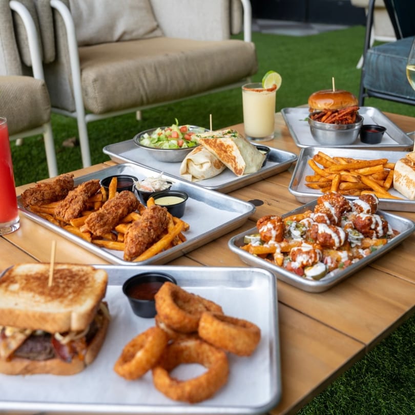 A wooden table laden with various bar-and-grill dishes and drinks on a patio with green turf.