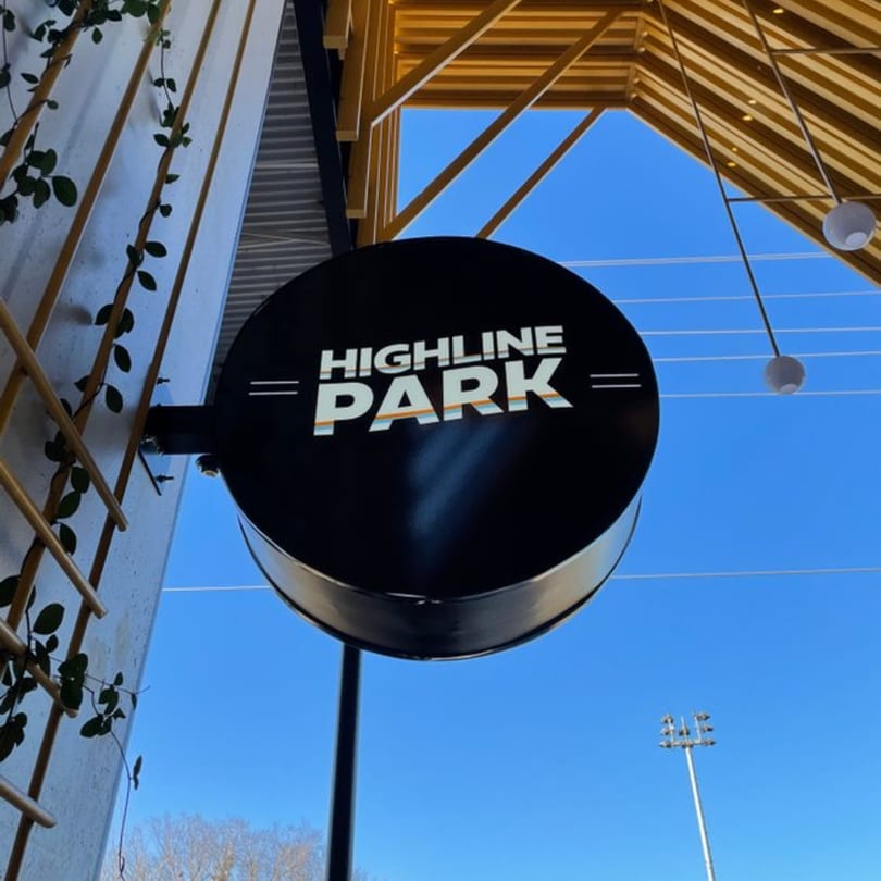 Highline Park restaurant sign, a black circle with white text, against a blue sky and wooden beams.