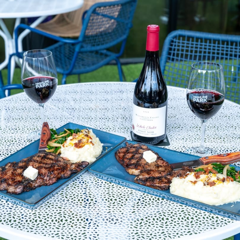 Two Highline Park steaks with mashed potatoes and wine on an outdoor patio table.