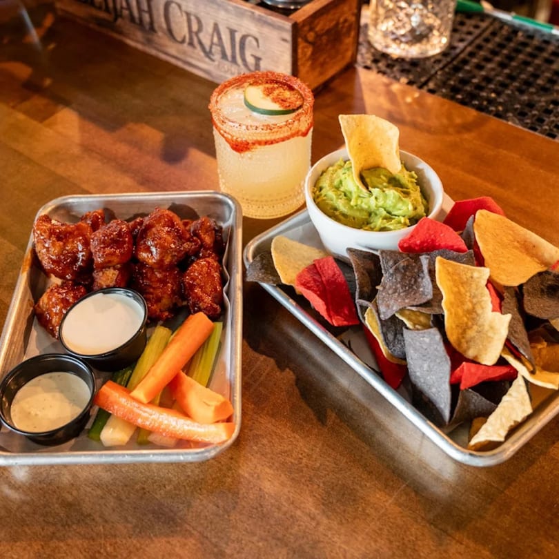 Highline Park bar-and-grill food: wings, guacamole, colorful tortilla chips, and a cocktail.