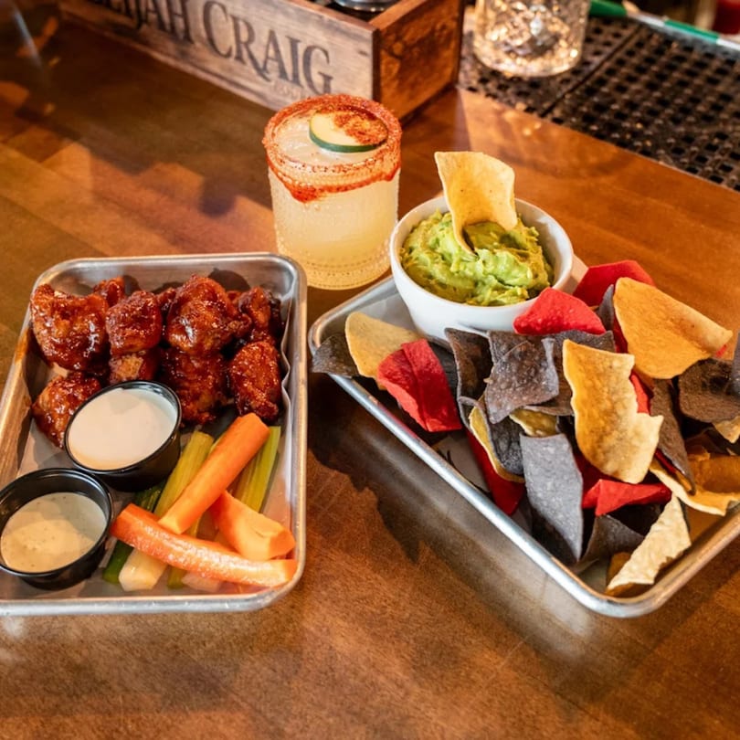 Highline Park's chicken wings, guacamole with colorful chips, and a cocktail on a wooden bar.