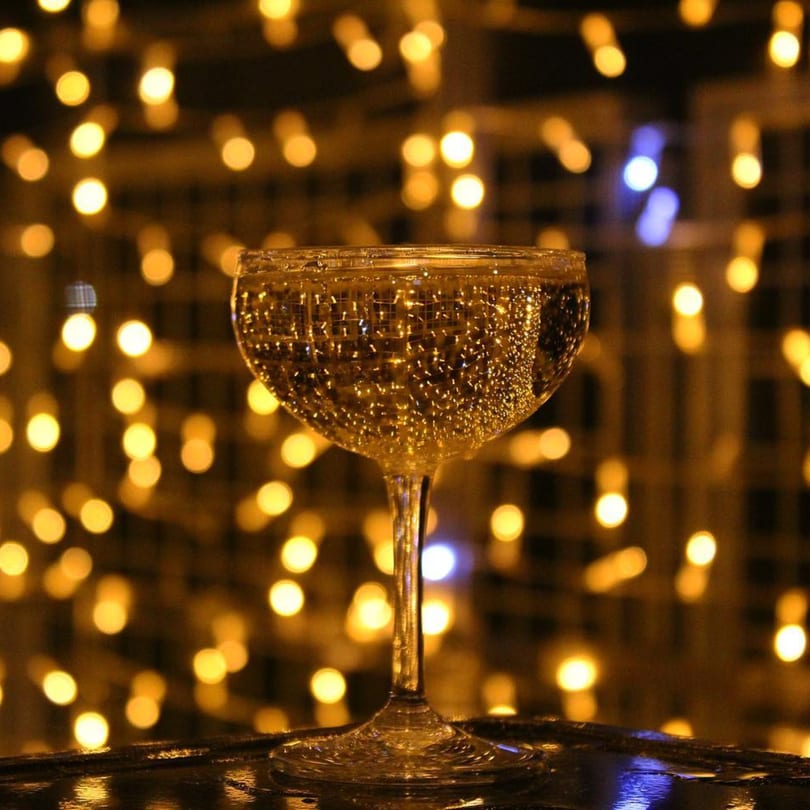 A coupe glass filled with a bubbly drink, set against a background of warm, blurred lights.