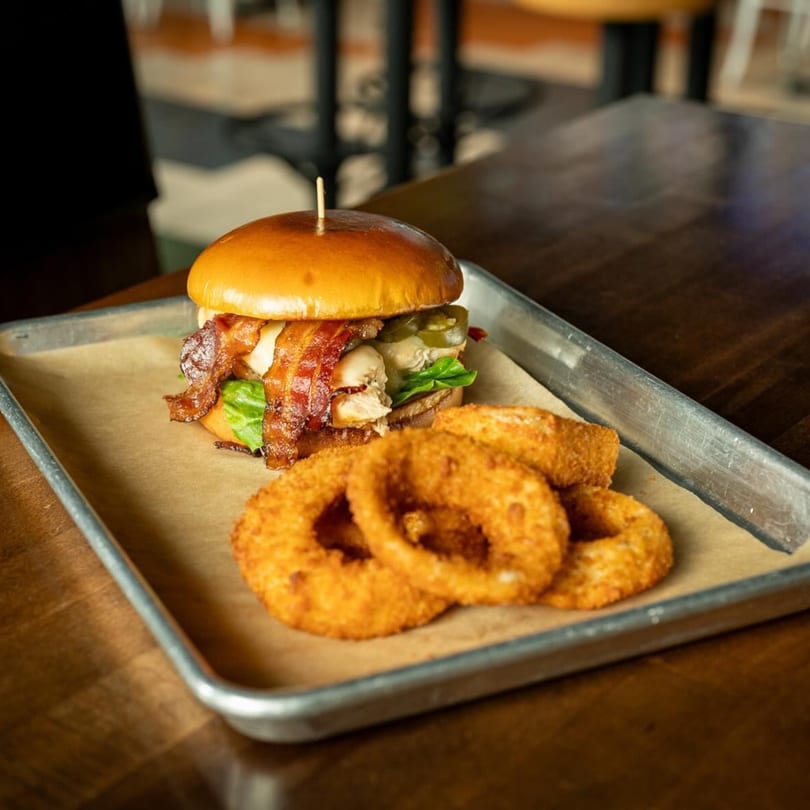 A Highline Park chicken sandwich with bacon and onion rings on a metal tray.