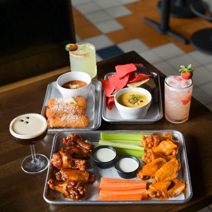 Highline Park bar-and-grill appetizers: wings, mozzarella sticks, chips with queso, and cocktails on a table.